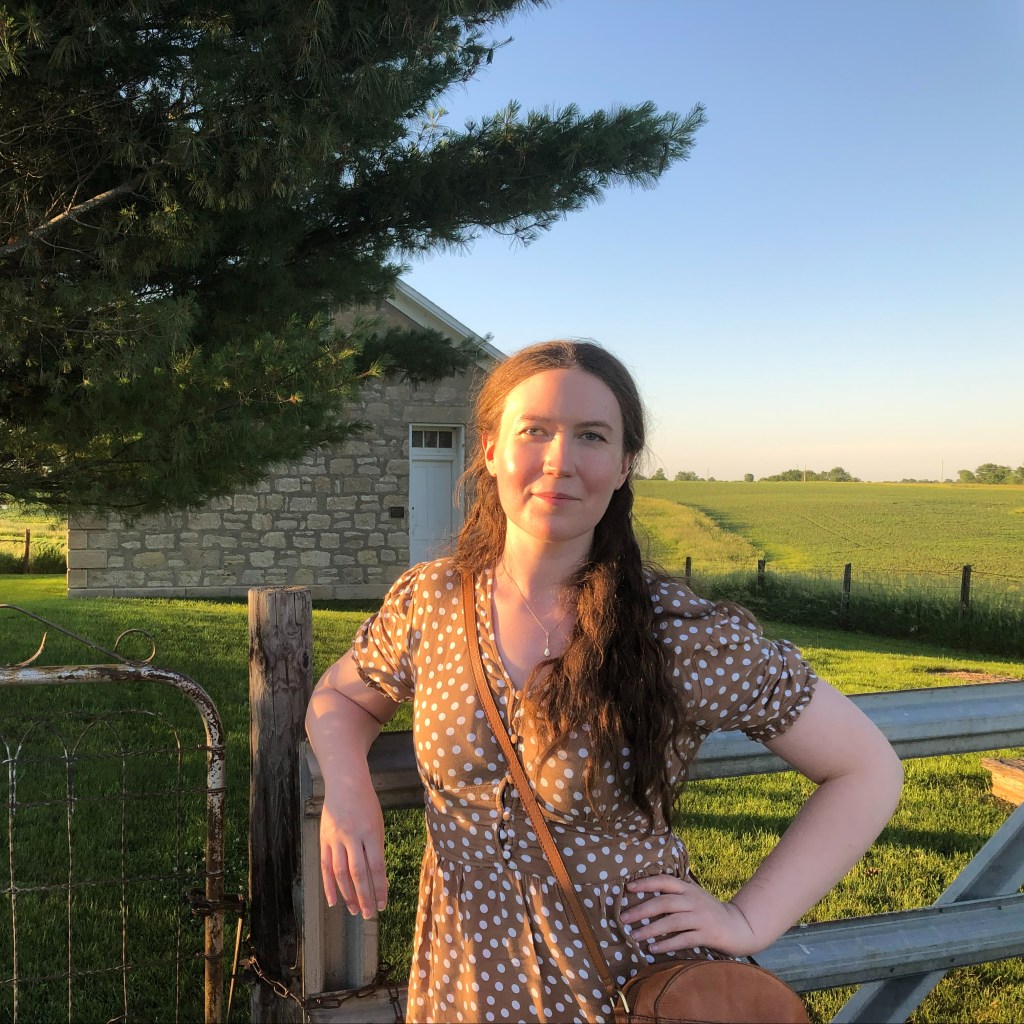 Alex in a brown polka dot dress leaning against a fence by a stone schoolhouse