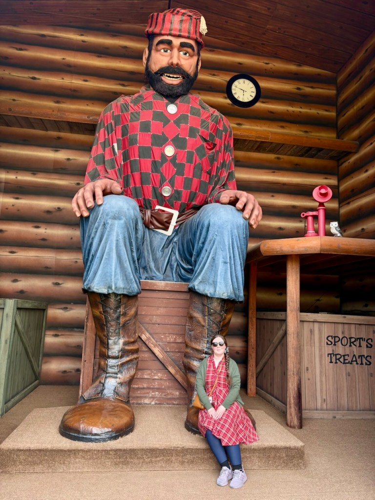 Alex in a red plaid dress sitting on the boot of a giant Paul Bunyan statue